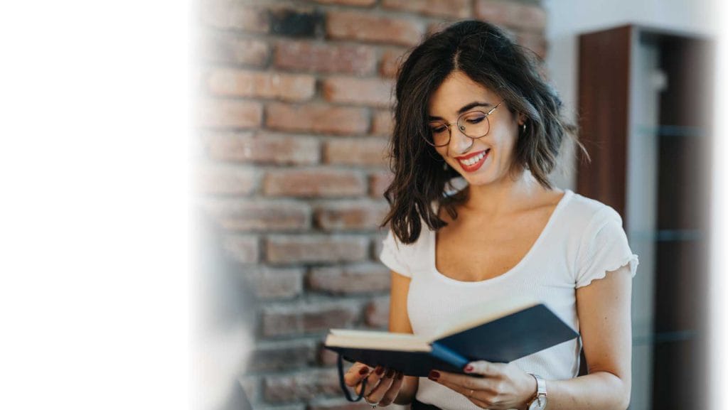 Smiling young woman in glasses enjoying reading a book in a cosy office